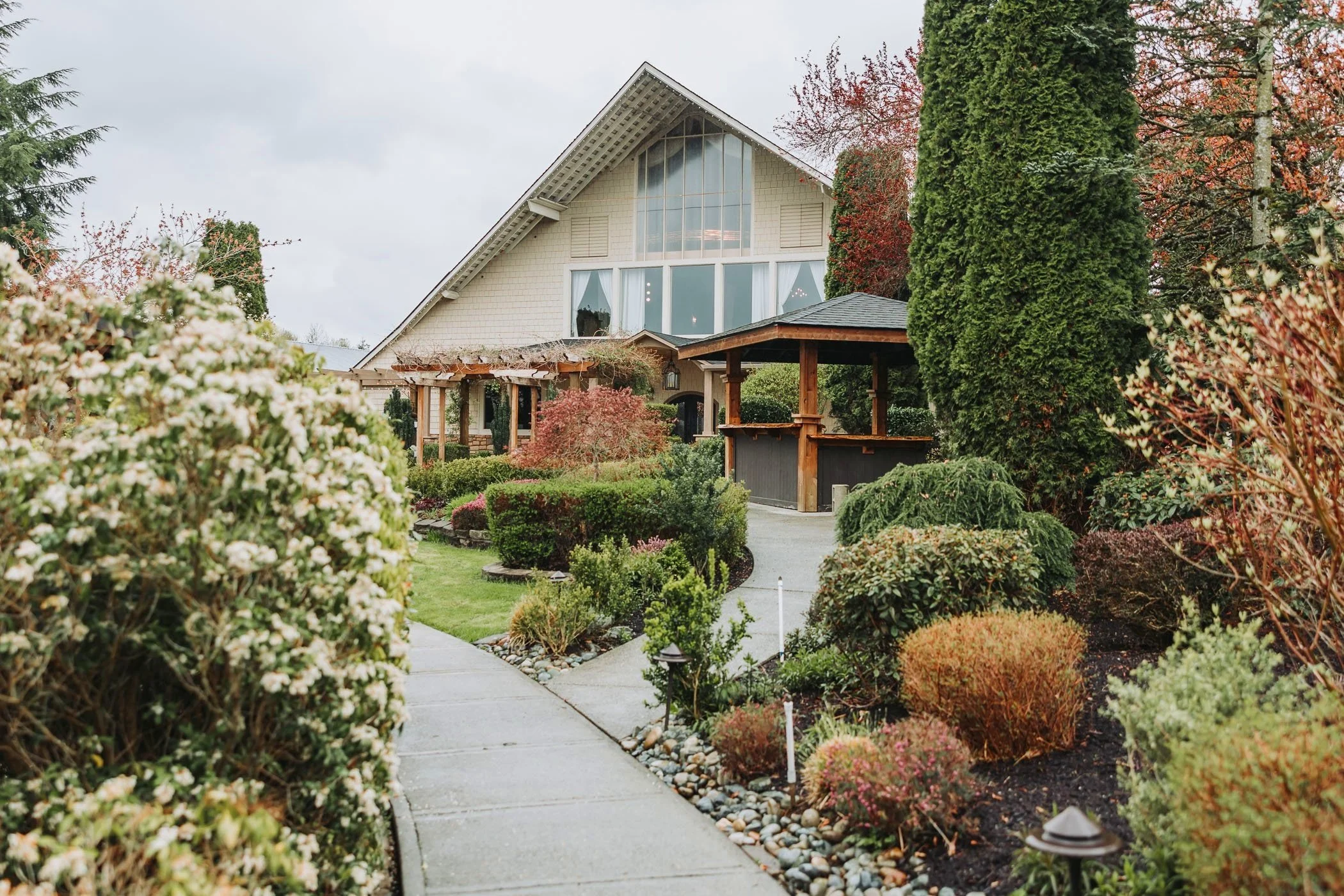 A house with large front windows, a sloped roof, and a well-maintained garden with various bushes, trees, and a pathway leading to the house in a suburban neighborhood on a cloudy day.